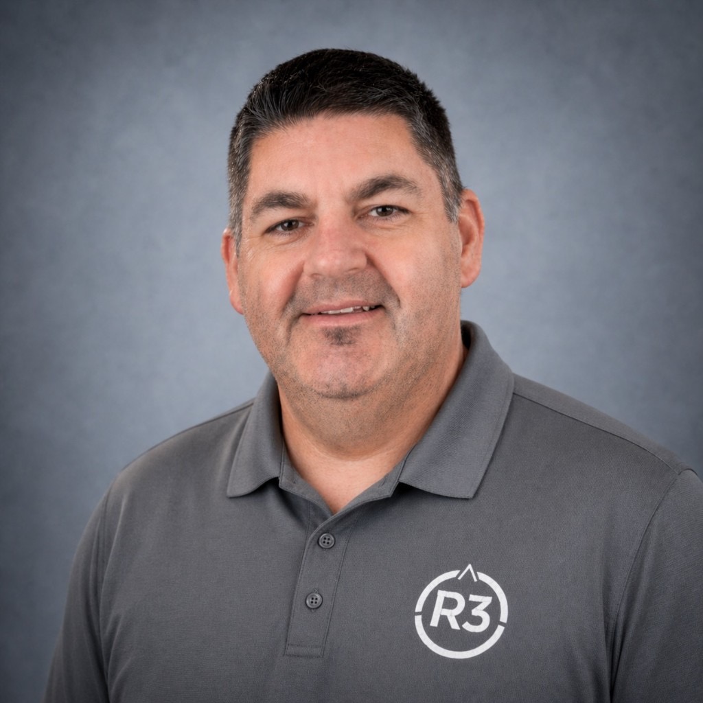 Joe Bourquin, professional head-and-shoulders portrait in a dark grey polo with the R3 logo, smiling at the camera against a blue-grey studio background.