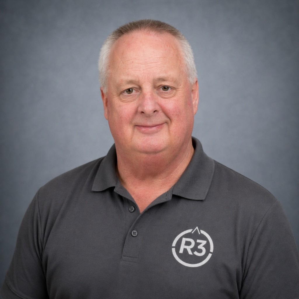 Marty Parker Sr., professional head-and-shoulders portrait in a dark charcoal polo with the R3 logo, smiling at the camera against a blue-grey studio background.