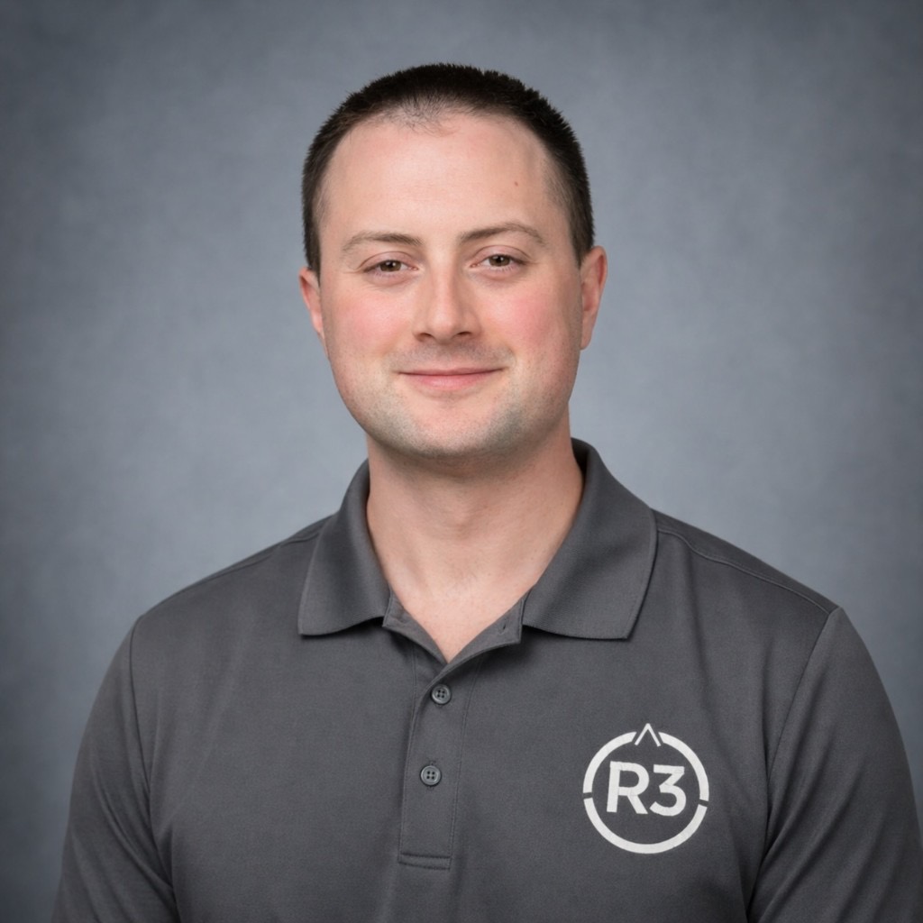 Marty Parker Jr., professional head-and-shoulders portrait in a dark grey polo with the R3 logo, smiling at the camera against a neutral grey studio background.